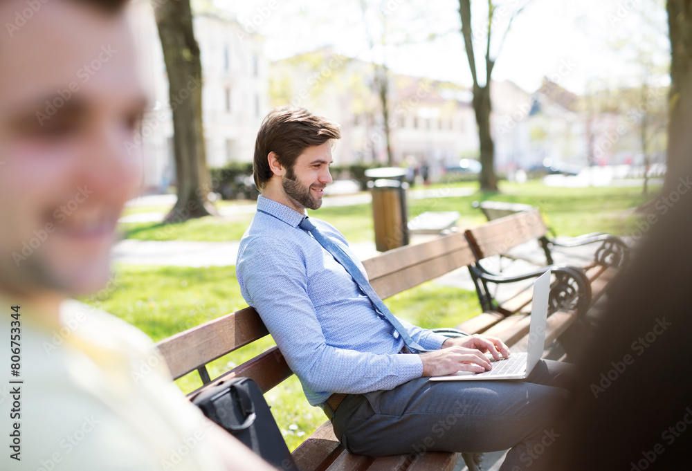 Handsome businessman sitting on city bench, typing on laptop. Working remotely from city park, waiting for business meeting. Manager smiling, outdoor in urban setting Handsome businessman sitting on city bench, typing on laptop. Working remotely from city park, waiting for business meeting. Manager smiling, outdoor in urban setting