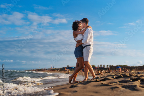 Young romantic couple - embracing lovingly on a sandy beach, enjoying a joyful moment at sunset by the sea - affectionate, warm.