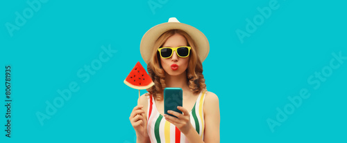 Summer portrait of happy young woman holding phone with juicy slice of watermelon on blue background