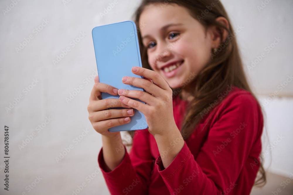 Modern smart mobile phone in the hands of a blurred smiling little child girl, a digital addicted kid browsing websites, playing online video games, watching bloggers or cartoons on internet. Modern smart mobile phone in the hands of a blurred smiling little child girl, a digital addicted kid browsing websites, playing online video games, watching bloggers or cartoons on internet.