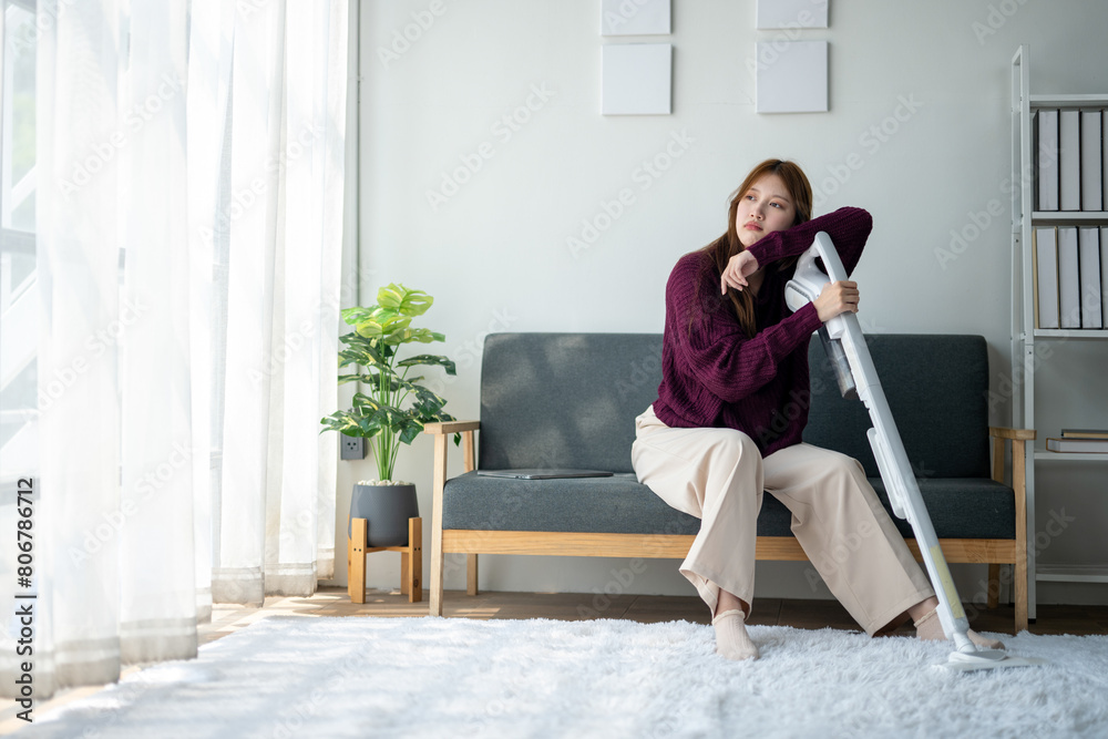 A woman sits on a couch with a vacuum cleaner in her hand. She looks tired and is leaning on the couch A woman sits on a couch with a vacuum cleaner in her hand. She looks tired and is leaning on the couch