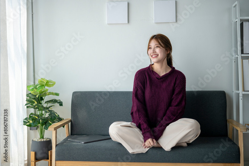 A woman is sitting on a couch with a laptop on her lap. She is wearing a purple sweater and has her hair in a ponytail. The room is decorated with a potted plant and a few books