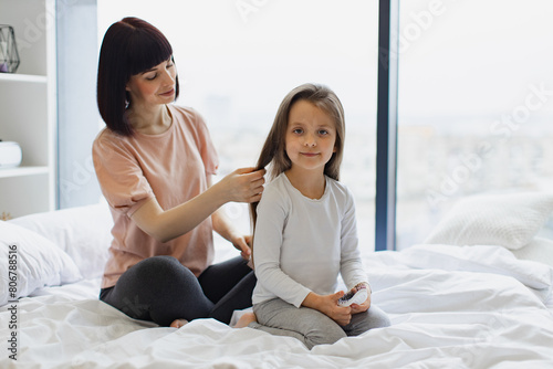 Charming little girl smiling while her cheerful mom brushing daughter's hair. Beautiful young mother combing hair to her daughter while sitting on the bed at home.