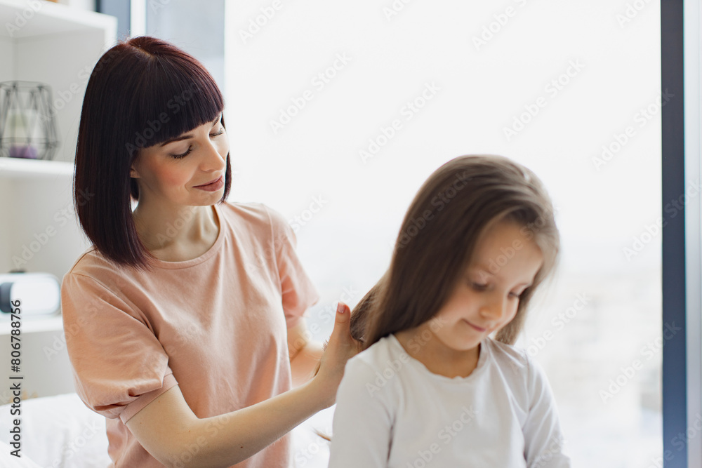 Charming little girl smiling while her cheerful mom brushing daughter's hair. Beautiful young mother combing hair to her daughter while sitting on the bed at home.