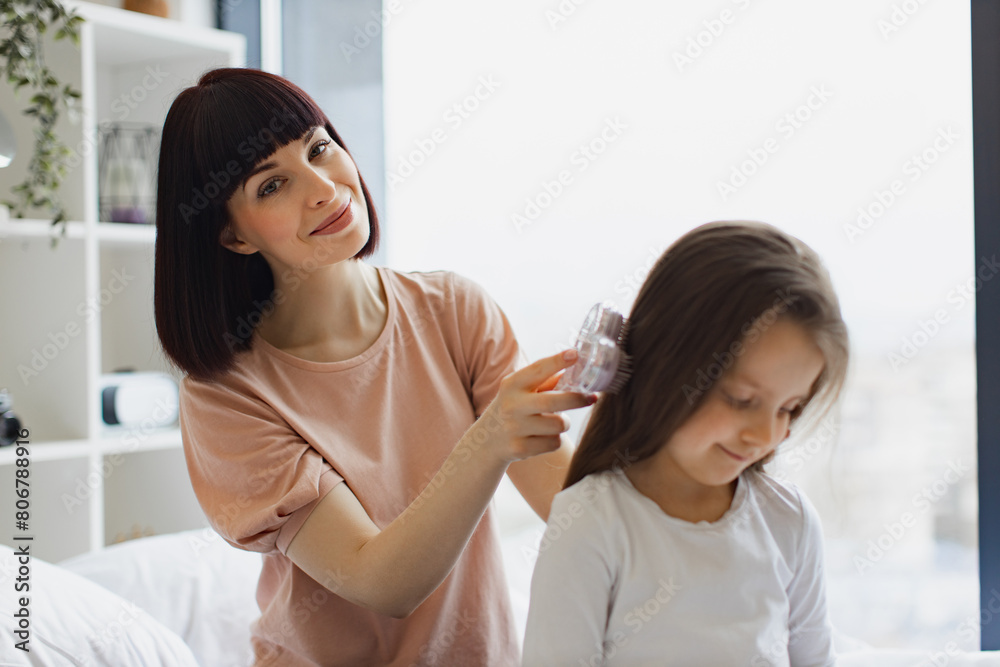Charming Caucasian mother combing her daughter's hair while sitting on bed near big panoramic window. Happy family spending weekend day home. Charming Caucasian mother combing her daughter's hair while sitting on bed near big panoramic window. Happy family spending weekend day home.