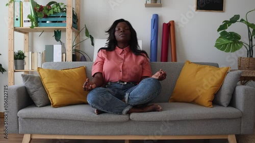 African American woman doing yoga on the couch at home relaxed meditating with her legs crossed and breathing deeply