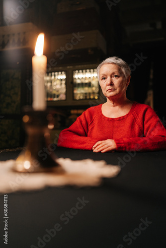 Vertical portrait of grey-haired adult woman receiving forecasting future during divination session sitting at table in dark room, by light of burning candle, looking at camera with serious expression