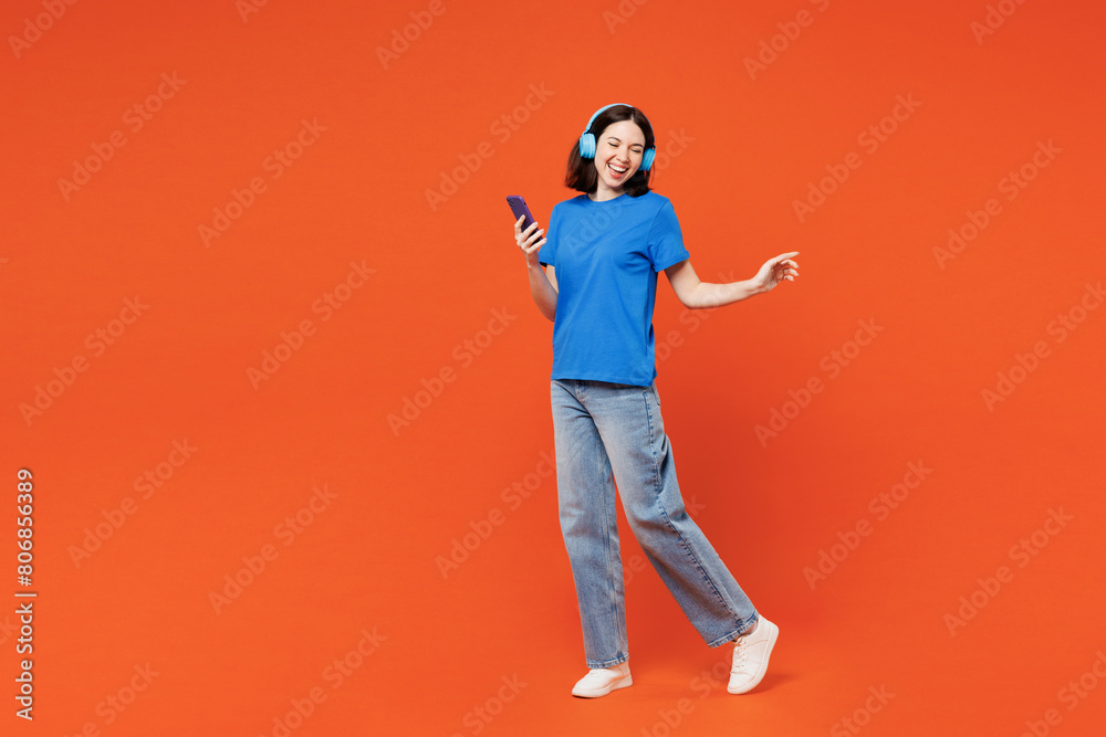 Full body young smiling happy woman she wear blue t-shirt casual clothes listen to music in headphones use mobile cell phone isolated on plain red orange background studio portrait. Lifestyle concept. Full body young smiling happy woman she wear blue t-shirt casual clothes listen to music in headphones use mobile cell phone isolated on plain red orange background studio portrait. Lifestyle concept.