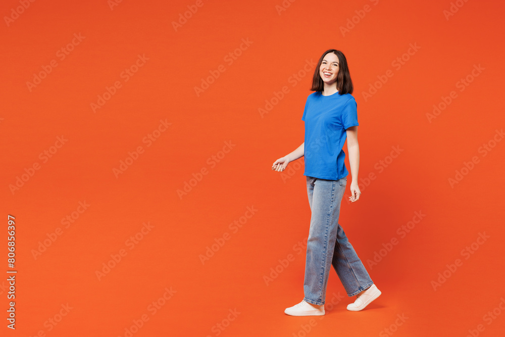 Full body side profile view young smiling happy woman she wears blue t-shirt casual clothes walk go look camera isolated on plain red orange color wall background studio portrait. Lifestyle concept. Full body side profile view young smiling happy woman she wears blue t-shirt casual clothes walk go look camera isolated on plain red orange color wall background studio portrait. Lifestyle concept.