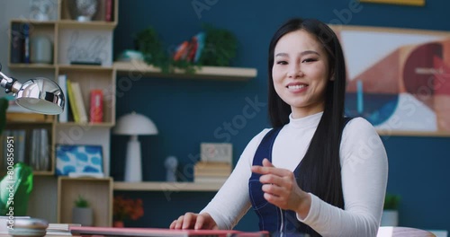 Cute Chinese female working or studying remotely. Pressing keys on keyboard of pink laptop. Shutting down computer. Closing deck lid with one hand. Looking directly at camera and showing heart symbol. photo