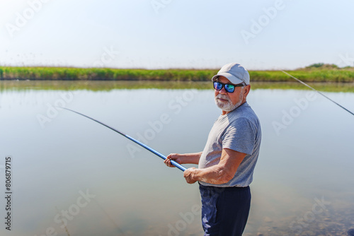 An elderly man in sunglasses and a cap stands on the bank of a river with a fishing rod. Rest and fishing on the pond.