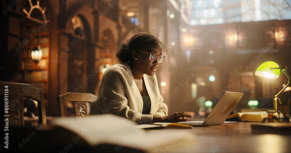 Focused African American College Student Studying in a Classic Library. Young Woman Using Laptop for Online Education, Surrounded by Books, Preparing for University Exams.