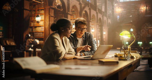 Engaged Multiethnic Students Collaborating on a Project in a Historic Library. Young Man and Woman Using Laptop, Discussing Academic Research Surrounded by Books in a Warmly Lit Study Environment.