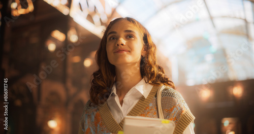 Confident Young Caucasian Female Student Carrying Books in a Grand Library. Smiling Woman in Casual Outfit, Looking Upwards with Optimism and Inspiration, Surrounded by Historical Architecture.