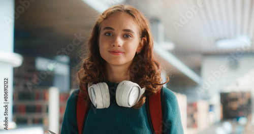 Young Female Student With Headphones Around Neck, Smiling In Modern School Corridor. Caucasian Teenager Carrying Books, Wearing Casual Teal Sweater and Red Backpack, Ready For Class. © Gorodenkoff