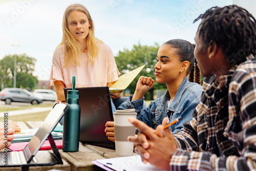 multiracial group of young students sitting together on a university campus bench with academic material studying or working on a class project.