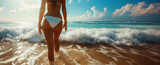 A slender woman in a white bikini walking on a sandy beach with waves crashing around her legs.