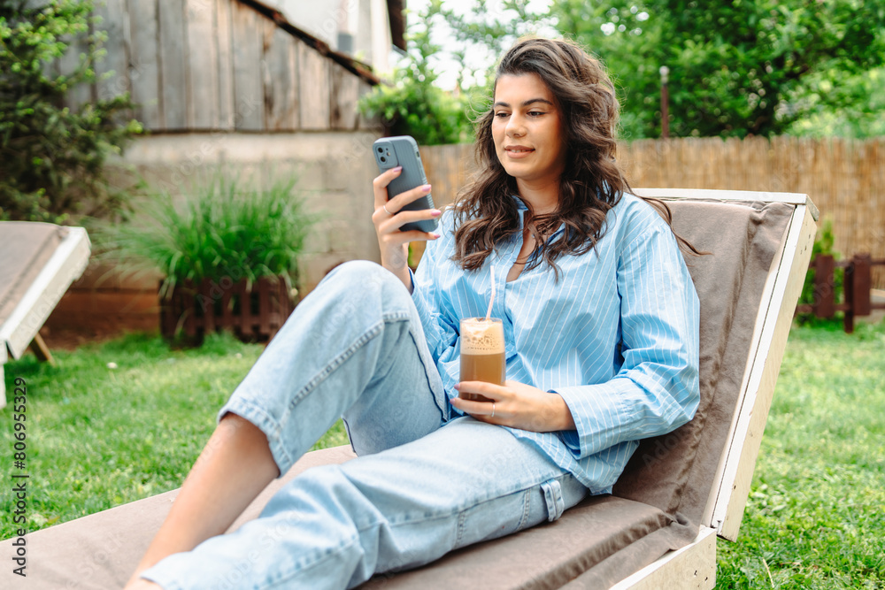 One young caucasian girl is relaxing on easy chair while drinking coffee and using mobile phone in her backyard One young caucasian girl is relaxing on easy chair while drinking coffee and using mobile phone in her backyard
