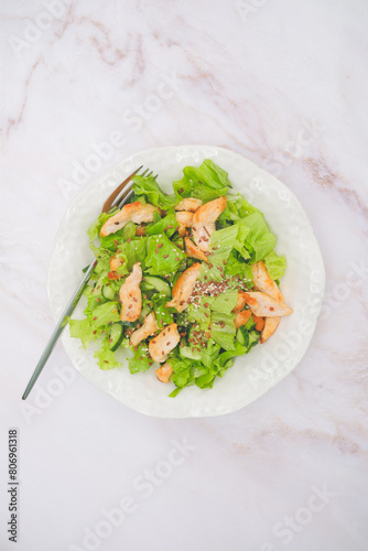Plate with healty salat - chicken, cucumber, lettuce leaves, flax seeds on white marble background. Top view.