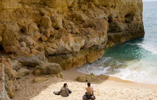 Back view of young girl and boy sitting on beach near Atlantic ocean and yellow rocks and talking. Sunny day. Summertime. Selective focus. Travel concept 