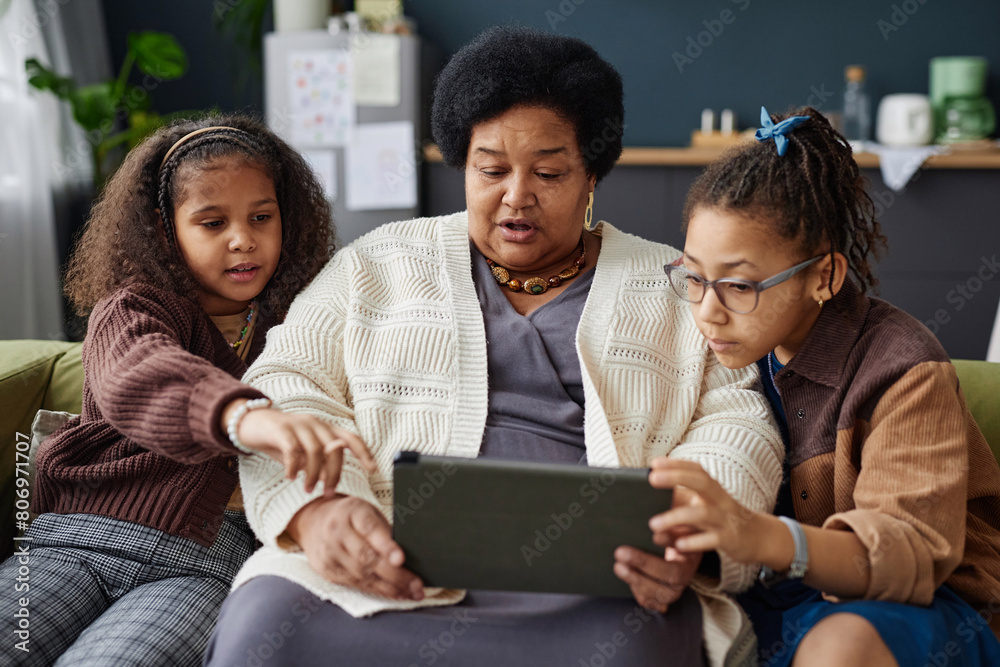 Obraz premium Front view portrait of Black senior woman using digital tablet at home with two children helping