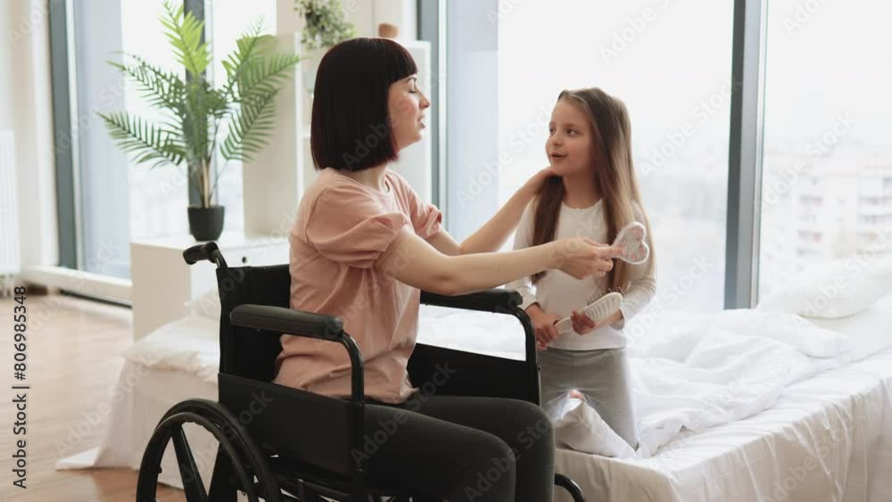 Yong mother wheelchair user brushing her little daughter's hair sitting near big panoramic window in bedroom. Happy Caucasian family having fun on bad on weekend morning.