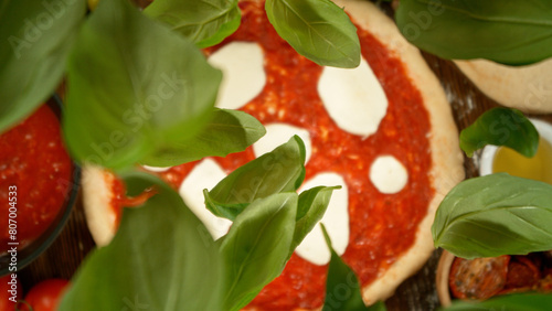 Super slow motion of falling fresh basil leaves on pizza dough with sugo, top down view