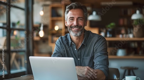 A smiling middle-aged businessman sits confidently at the table. The laptop is open in front of you. This is a testament to the power of technology. Bringing people together to achieve shared goals