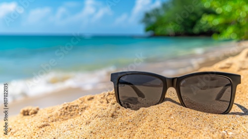  A pair of black sunglasses rests atop a sandy beach, near a tranquil body of water and trees in the background