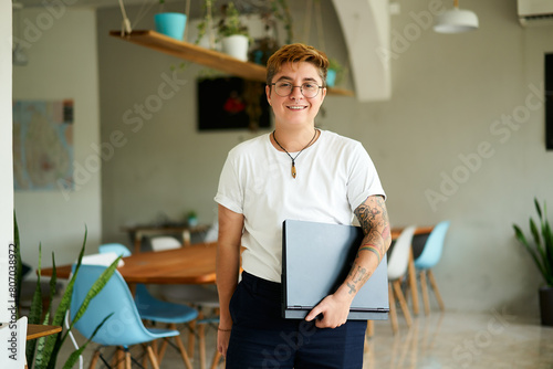 Confident transgender person with tattoos holds laptop, smiles in modern eco office. Inclusive LGBTQ work environment, gender identity acceptance at tech startup. Casual corporate attire. photo