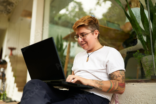 Tattooed Gen Z transgender person works on laptop in cozy office space, displays casual, inclusive workspace. Focused yet comfortable, represents modern work-life balance, LGBTQ-friendly environment.