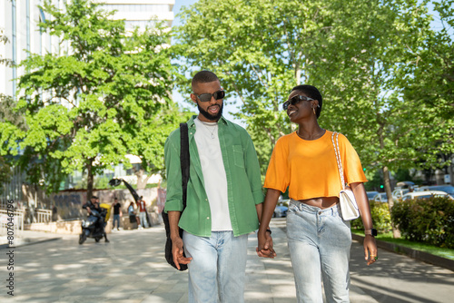 Happy pleased african american couple strolls through city, chatting smiling enjoy time together on date. Contented black girl going in sidewalk downtown holds hands with guy talks pleasant dialogue.