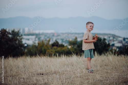 happy little boy stands on hill, family trip on summer day at sunset. child smiles slyly and plays with wooden airplane. Beautiful view sea and city, holidays with children, vacations, family leisure