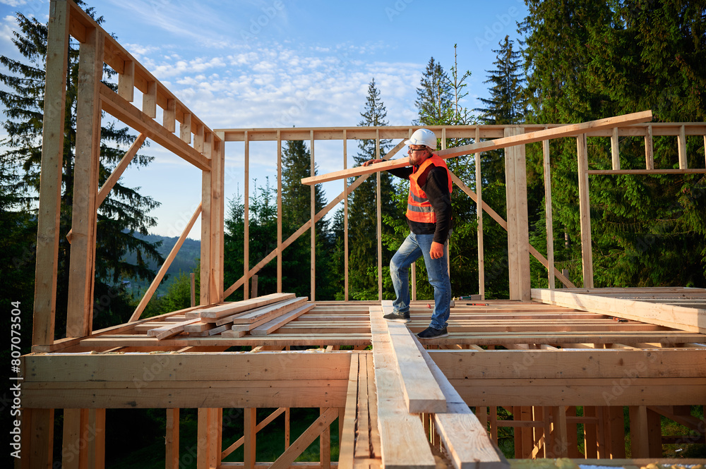 Carpenter building wooden-framed house near the forest. Man holds large board, dressed in work clothes and helmet. Concept of contemporary and eco-friendly construction. Carpenter building wooden-framed house near the forest. Man holds large board, dressed in work clothes and helmet. Concept of contemporary and eco-friendly construction.