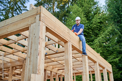 Joiner erecting wooden, two-story, frame-built dwelling adjacent to the forest. Bearded, bespectacled man hammering nail with hard hat secured. The principle of present-day ecological construction.