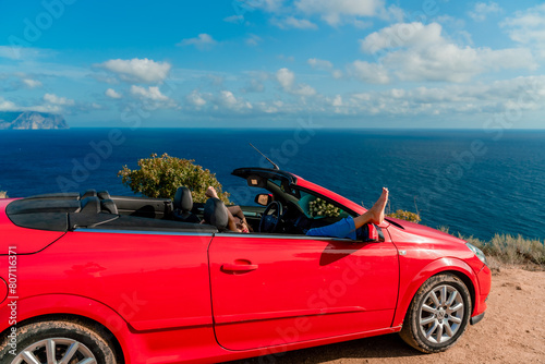A woman is laying on her feet in a red car, looking out the window at the ocean © svetograph