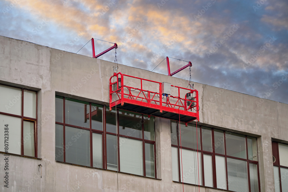 Red suspended scaffolding platform on facade of building under renovation. Plastering works at height, high-altitude plaster works on facade. Building renovation. Mobile scaffolding platform