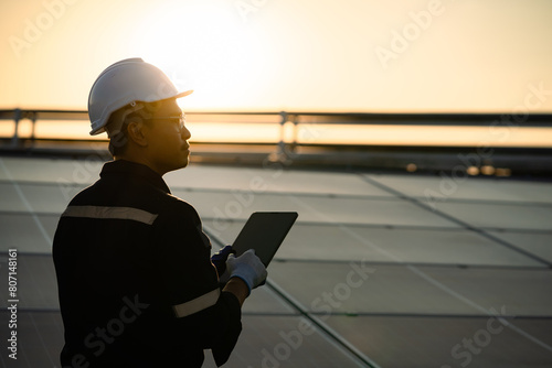 A man wearing a hard hat and safety glasses is holding a tablet in his hand. He is looking up at the sky, possibly at the sun. Concept: Renewable energy, clean technology, electric energy