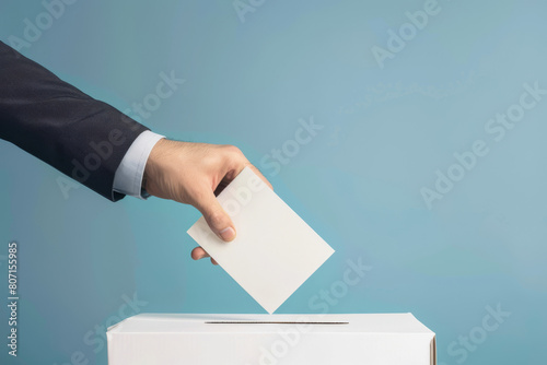 Close-up of a person's hand in a suit sleeve, inserting a blank voting slip into a white ballot box against a soft blue background, conveying the act of voting in democratic elections photo