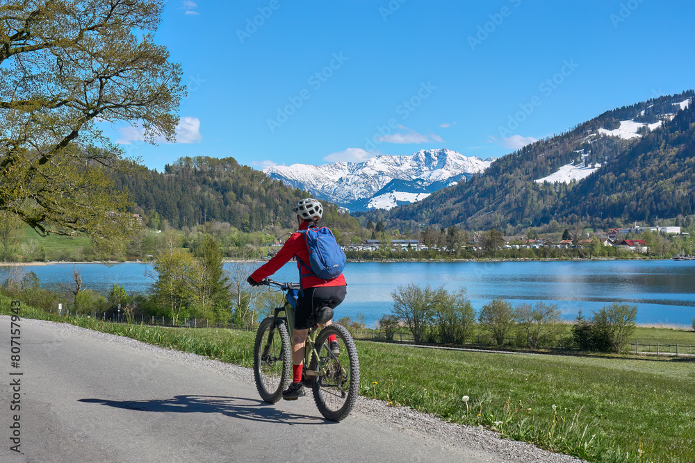 Obraz premium nice senior woman riding her electric mountain bike in springtime at lake Alpsee near Immenstadt, with snowcapped High Algaeu mountains in Background,Allgau,Bavaria, Germany