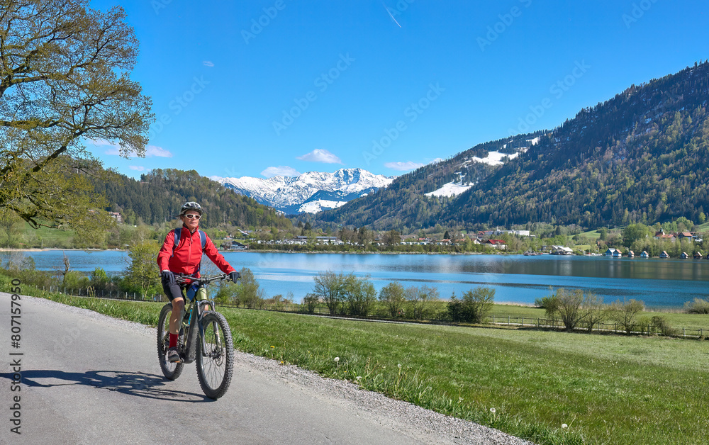 Obraz premium nice senior woman riding her electric mountain bike in springtime at lake Alpsee near Immenstadt, with snowcapped High Algaeu mountains in Background,Allgau,Bavaria, Germany