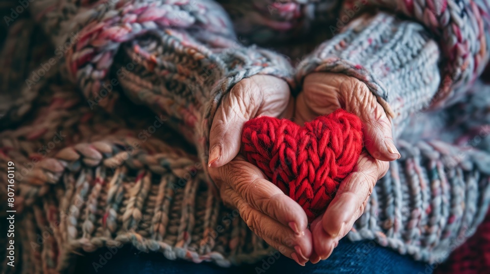 An elderly person is holding a red heart-shaped ornament in their hands. The hands and the ornament are in focus, the rest of the image is blurry.
