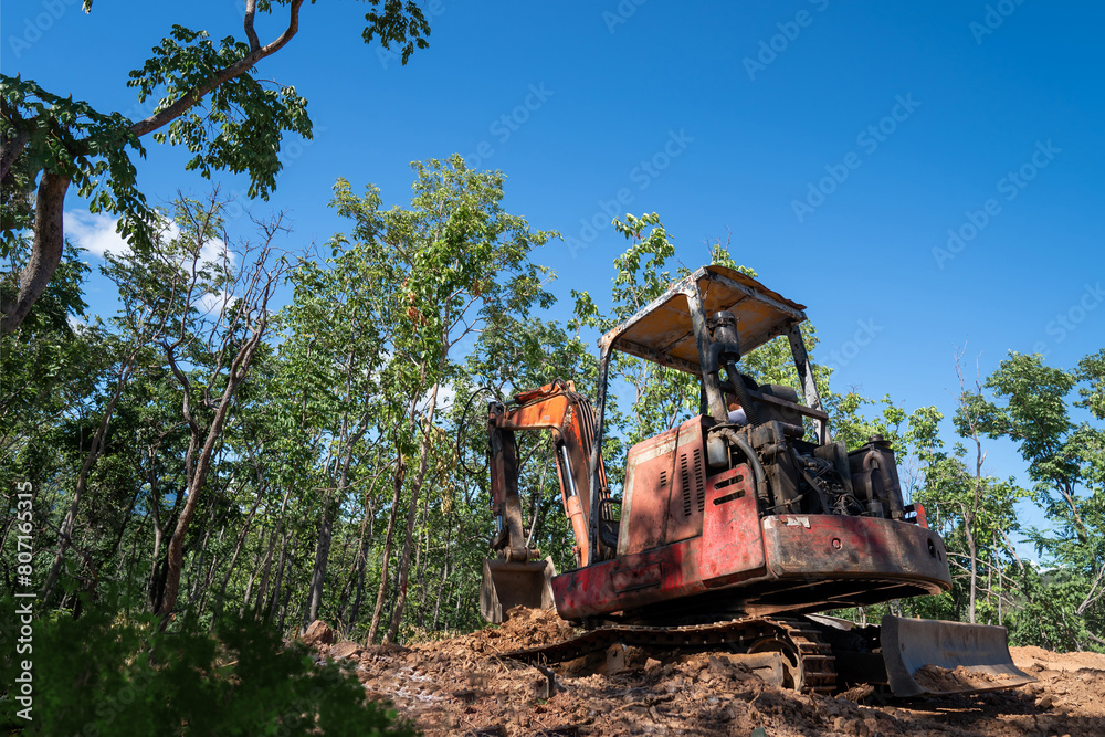 Hydraulic excavator is digging soil at a construction site, surrounded by lot of trees, under clear sky. Clawer backhoe parked under tree shade. Hydraulic excavator is digging soil at a construction site, surrounded by lot of trees, under clear sky. Clawer backhoe parked under tree shade.
