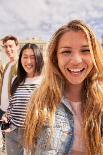 Vertical. Focus on cheerful young blonde woman looking at camera on street group multiracial friends in background. Smiling people together posing portrait. Gen z colleagues sightseeing on vacation