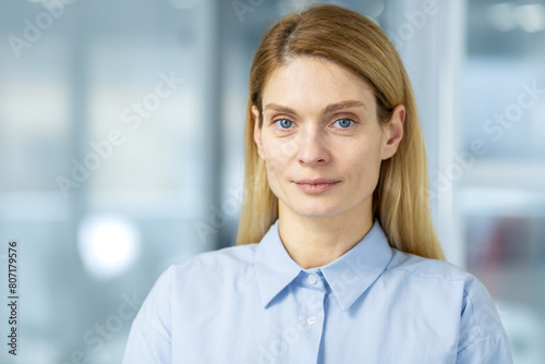 Close-up of a professional woman with blond hair and blue eyes, confidently smiling in a modern office. Depicts leadership and competence in a corporate environment.