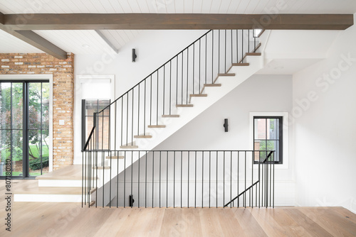 A beautiful staircase with wood flooring and stair treads, wrought iron railings, wood beams across the ceiling with shiplap, and a brick wall around a black framed window.