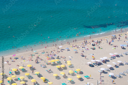 Beautiful top view of blue sea ocean water and beach shore with lots many of people tourists under white yellow umbrellas in summertime and bright sunlight sun.Tourism destination,vacation,holiday