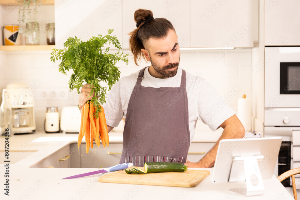 A man is holding a bunch of carrots and preparing to cut them on a cutting board. He is wearing an apron and has a serious expression on his face A man is holding a bunch of carrots and preparing to cut them on a cutting board. He is wearing an apron and has a serious expression on his face