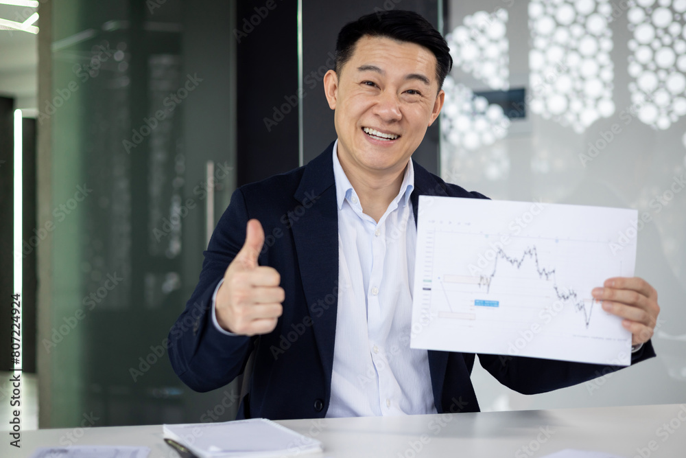 Smiling Asian businessman in a suit giving a thumbs up while holding a financial chart during a video call in a modern office setting. Smiling Asian businessman in a suit giving a thumbs up while holding a financial chart during a video call in a modern office setting.