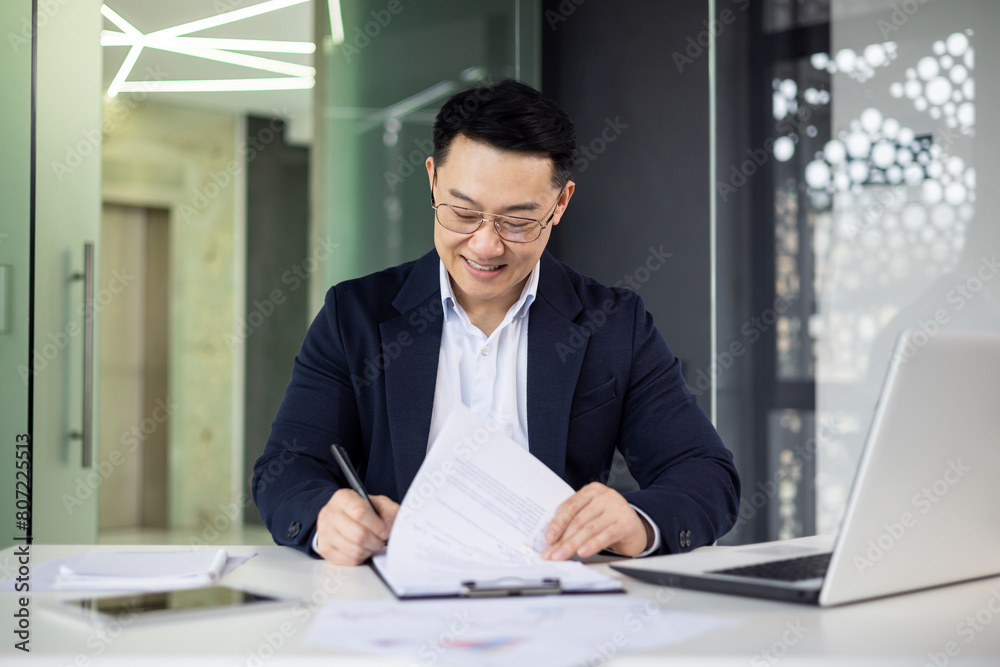 Smiling Asian businessman in a formal suit reviews documents while working on a laptop in a contemporary office setting with natural lighting.